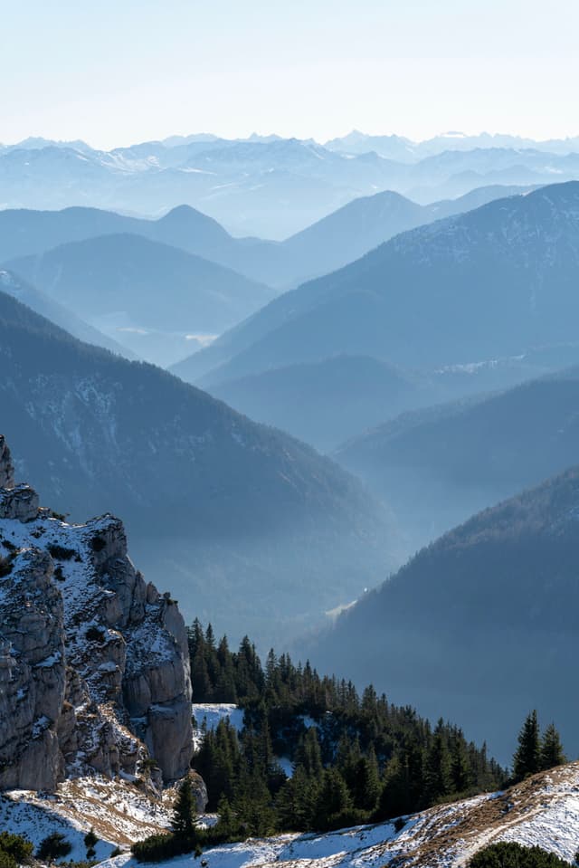 Snow-dusted rocky slope and evergreen forest descending into a series of hazy blue mountain ridges fading into the distance