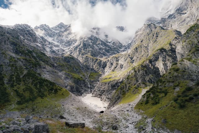 Steep rocky mountains with patches of snow and green slopes rise around a narrow valley under a cloudy sky
