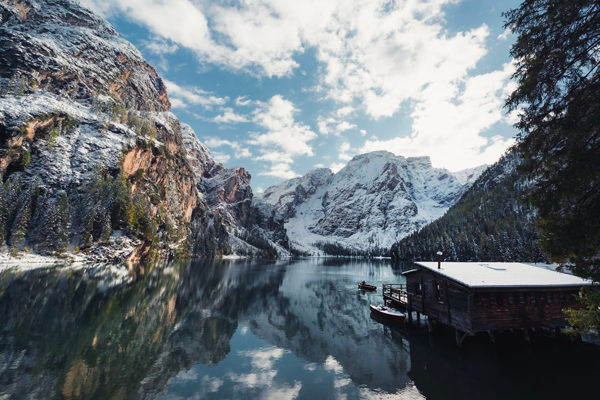 Snow-dusted mountains reflected in a calm alpine lake beside a small wooden boathouse under a partly cloudy sky