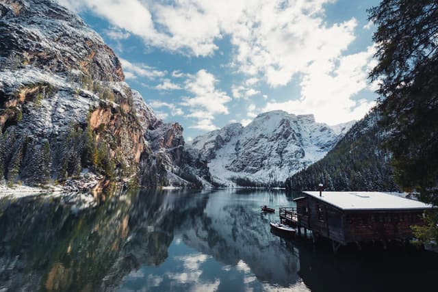 Snow-dusted mountains reflected in a calm alpine lake beside a small wooden boathouse under a partly cloudy sky