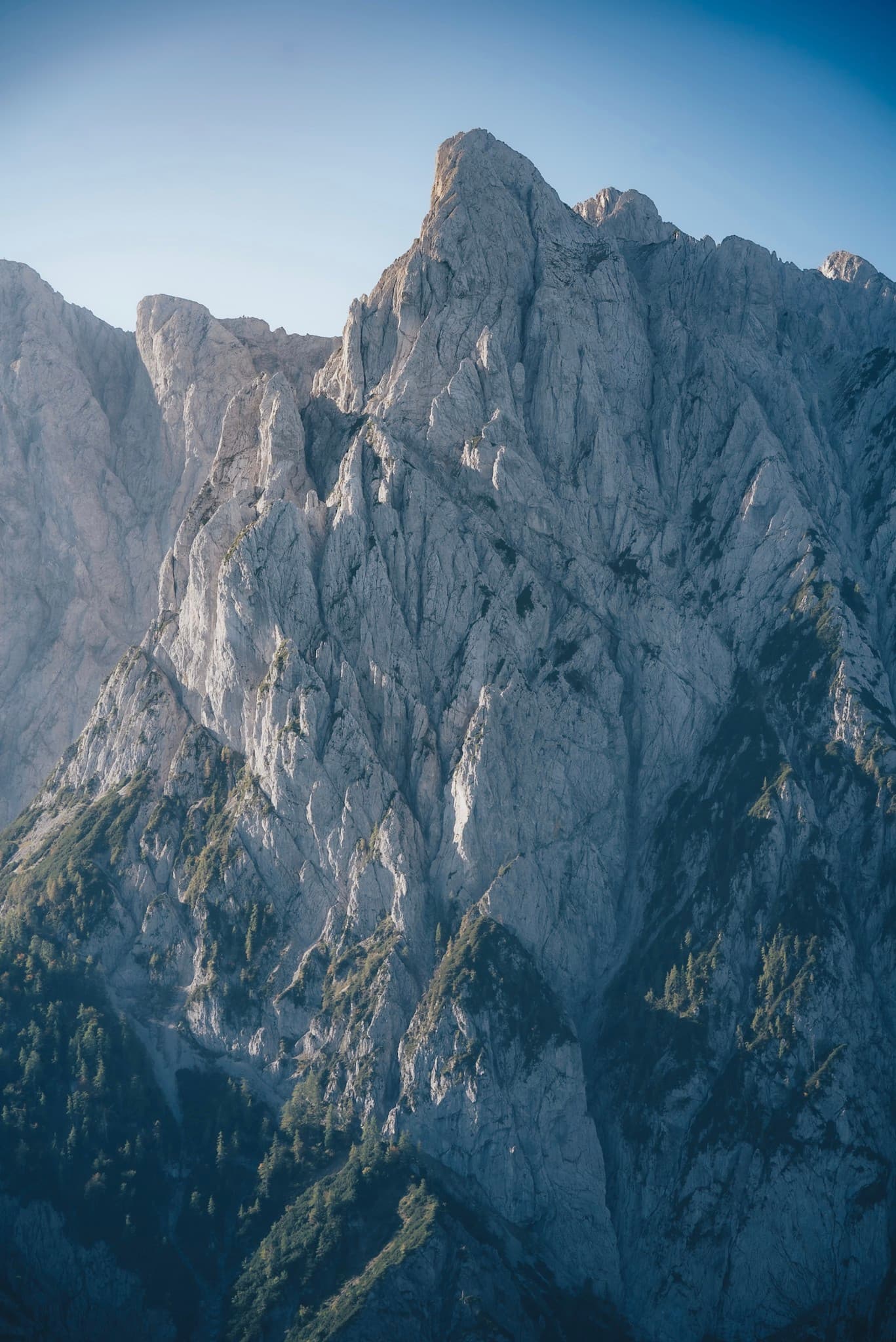 Steep jagged mountain cliffs rising sharply with patches of green forest below under a clear blue sky