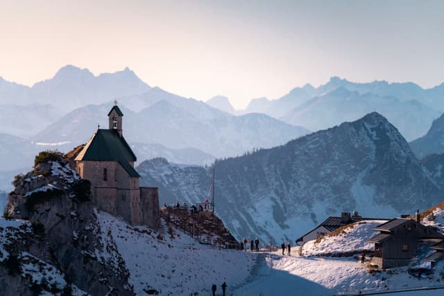 Small chapel perched on a snowy mountain ridge with distant layered peaks and tiny hikers walking along the path below