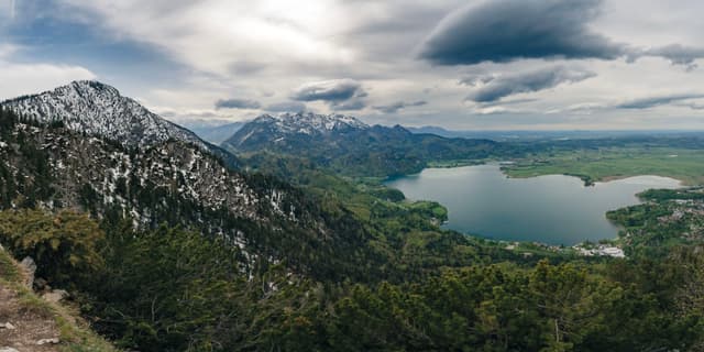 A panoramic view of a mountainous landscape with snow-capped peaks, a large lake, and dense forests under a cloudy sky