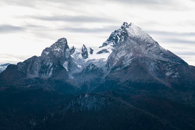 Snow-dusted jagged mountain peaks rising above dark forested slopes under a cloudy sky