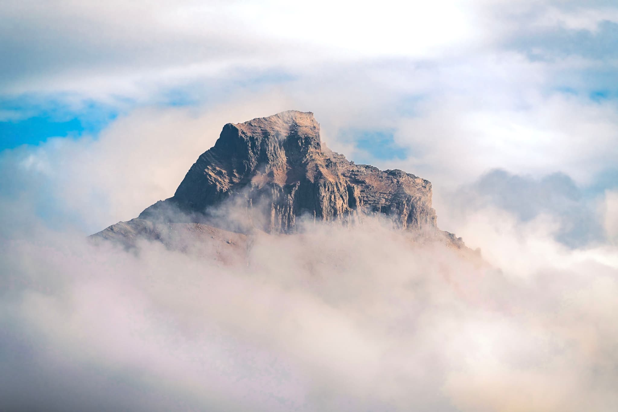 Jagged mountain peak emerging through a thick layer of clouds under a bright sky