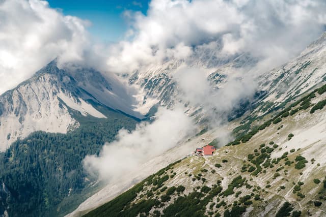 High alpine slopes with scattered trees and a solitary red mountain hut nestled among rocky ridges beneath drifting clouds