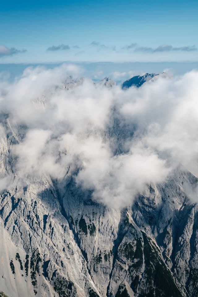 Jagged rocky mountain peaks partially obscured by soft white clouds under a clear blue sky