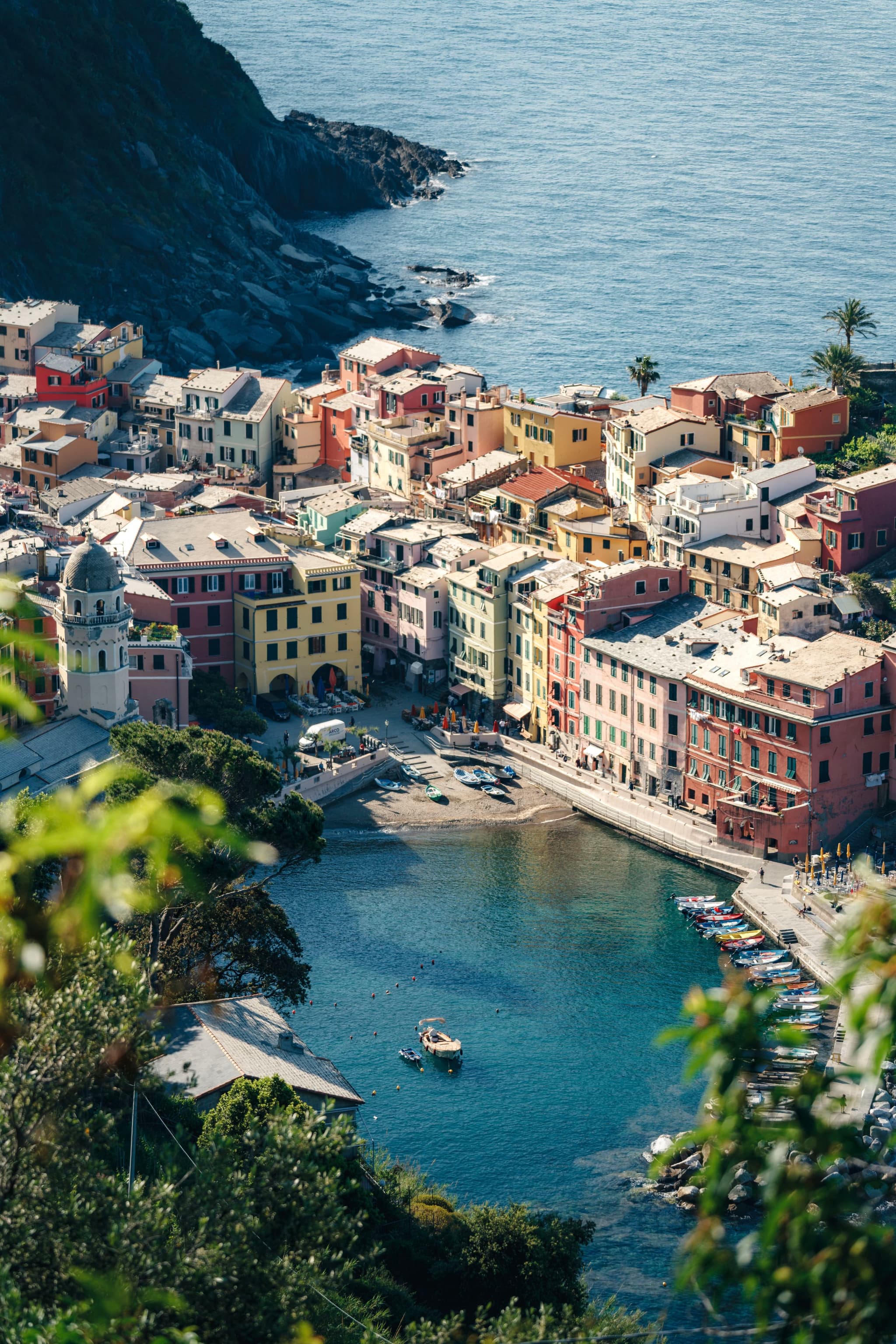 A coastal village with colorful buildings clustered around a small harbor, surrounded by lush greenery and a rocky shoreline