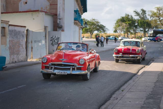 Two vintage red convertibles driving down a sunny urban street with pedestrians and trees in the background