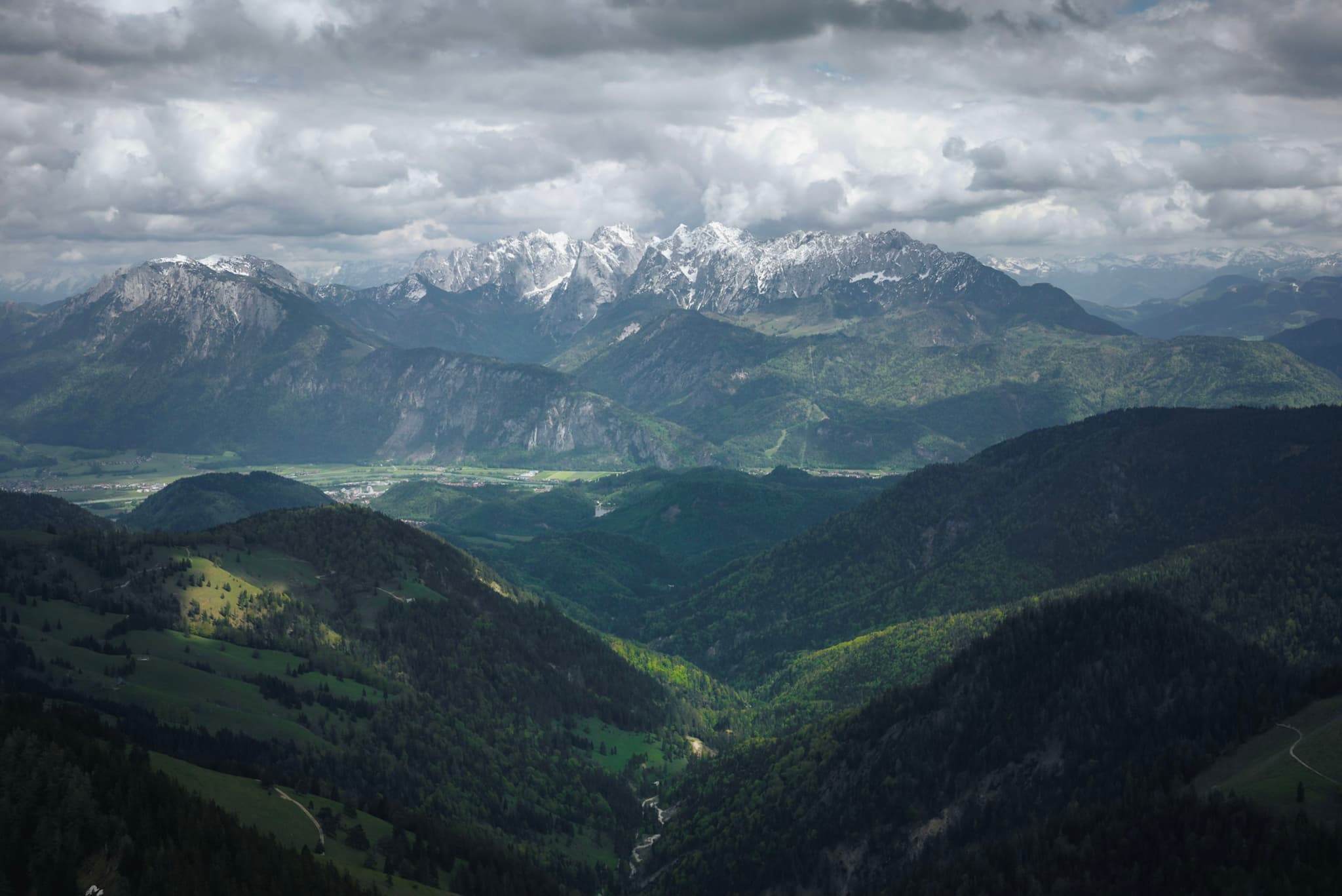 Layered green valleys leading toward distant snow-dusted mountains under heavy, dramatic clouds