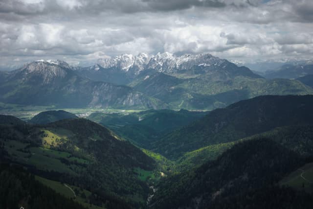 Layered green valleys leading toward distant snow-dusted mountains under heavy, dramatic clouds