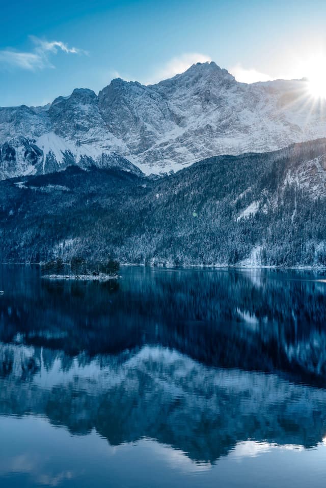 A serene mountain landscape with snow-capped peaks, a clear blue sky, and a sunburst effect. The mountains are reflected in a calm lake surrounded by forested hills