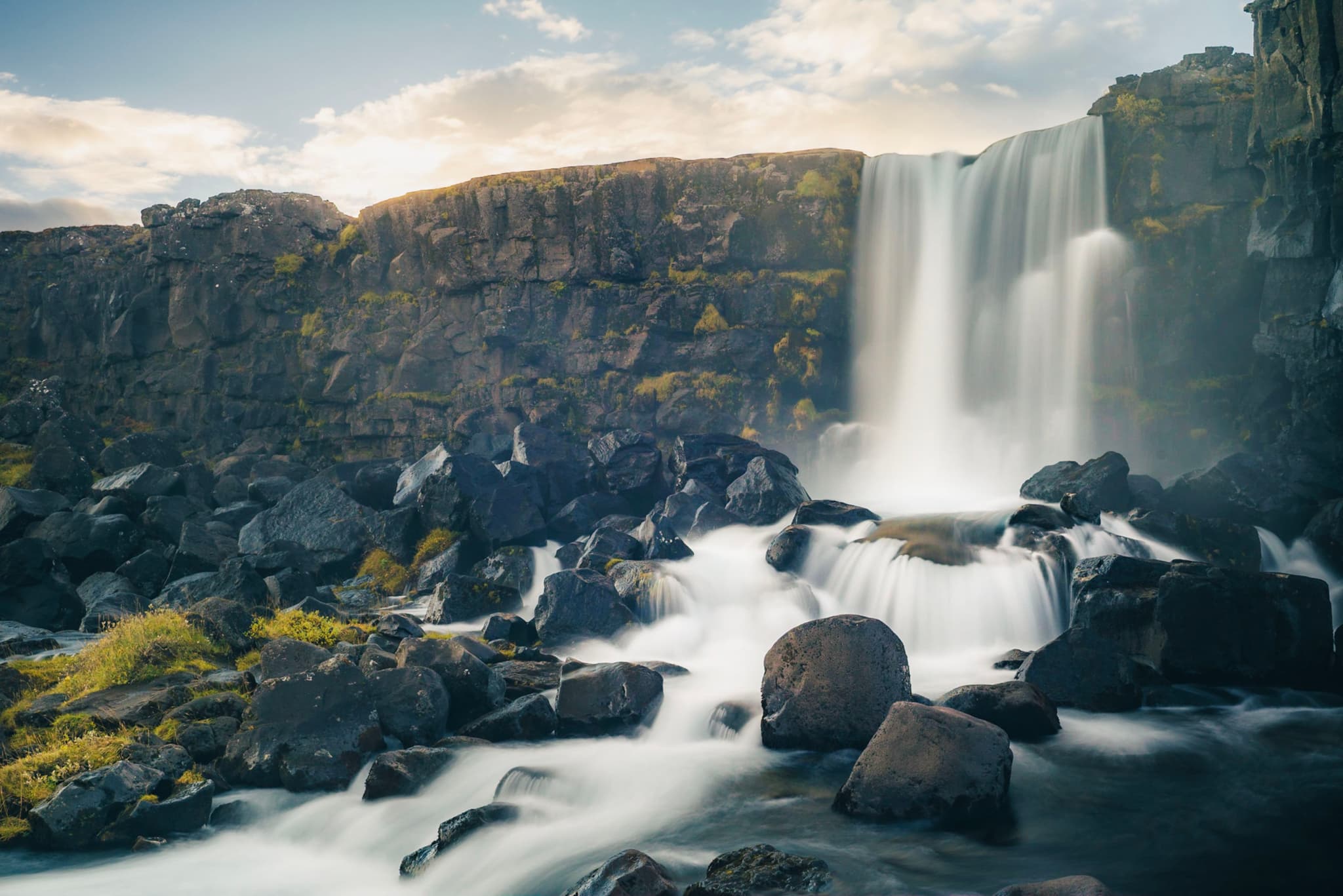 Tall waterfall pouring over a rocky cliff into a stream flowing around large dark boulders under a bright cloudy sky