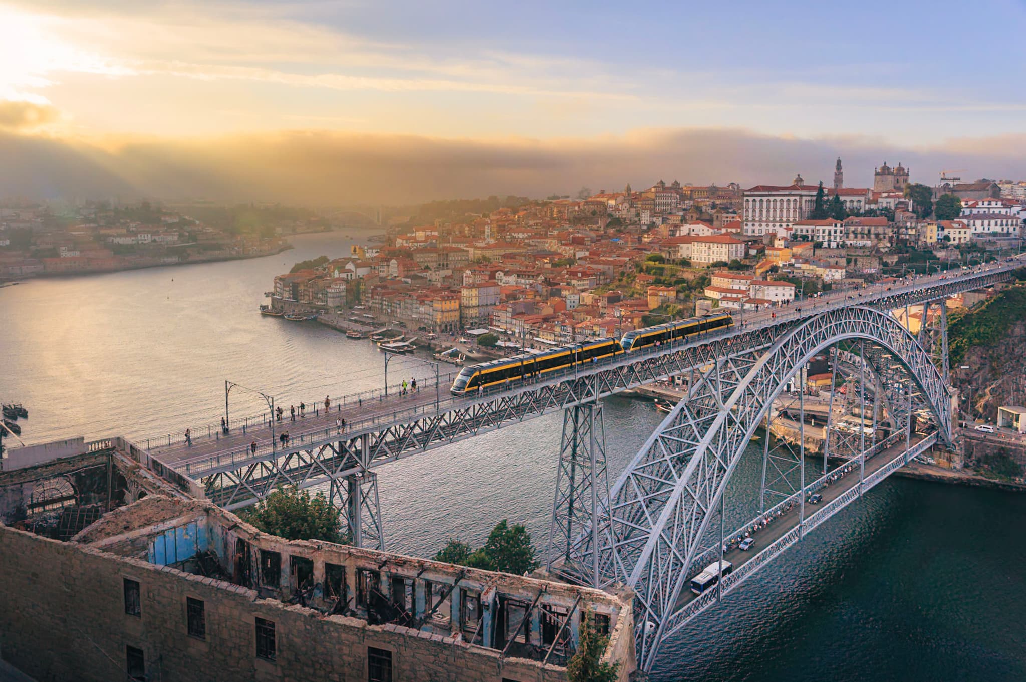 Large arched metal bridge spanning a wide river with a historic riverside city and hazy sunset in the background
