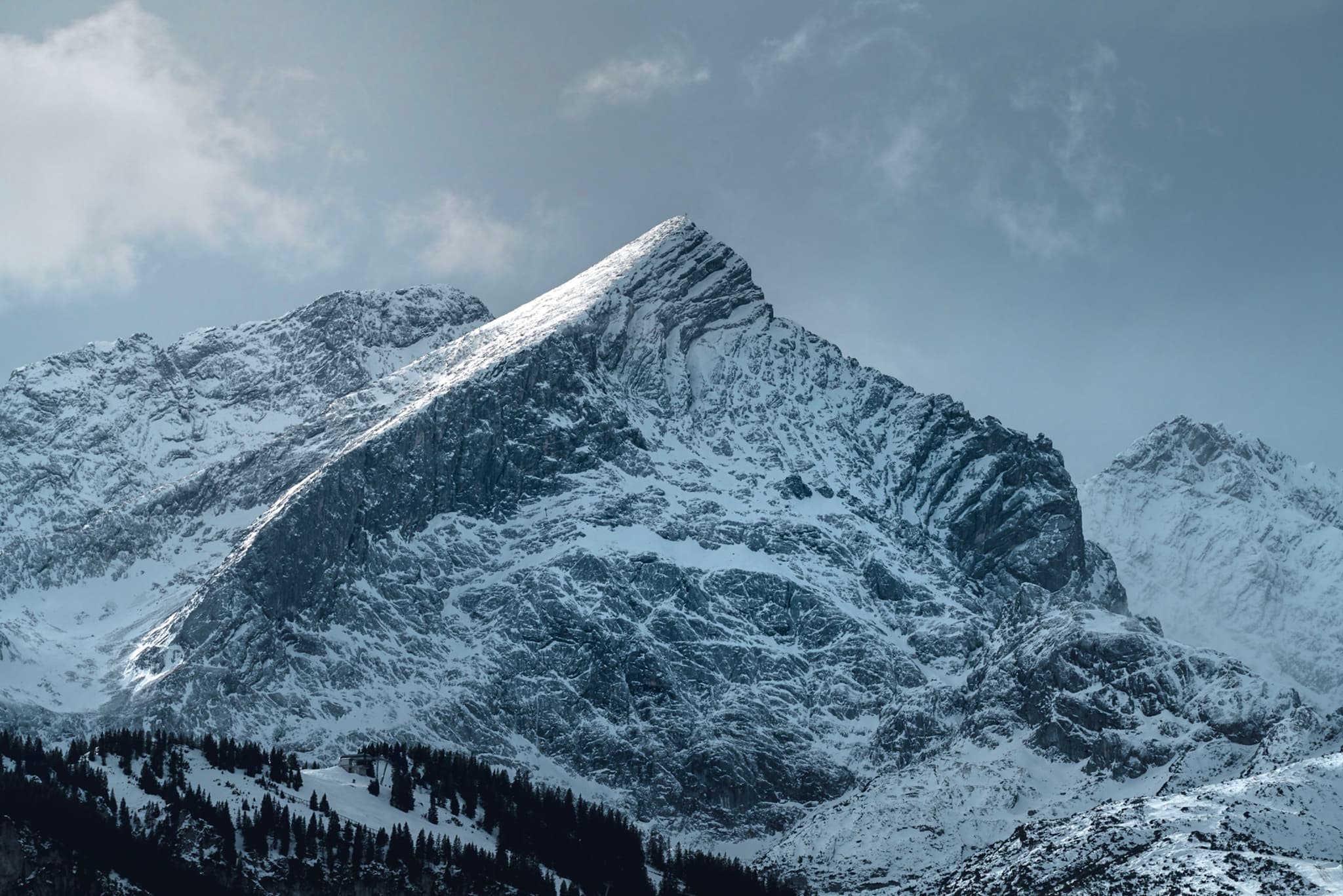Jagged snow-covered mountain peak under a moody, overcast sky with dark forested slopes below