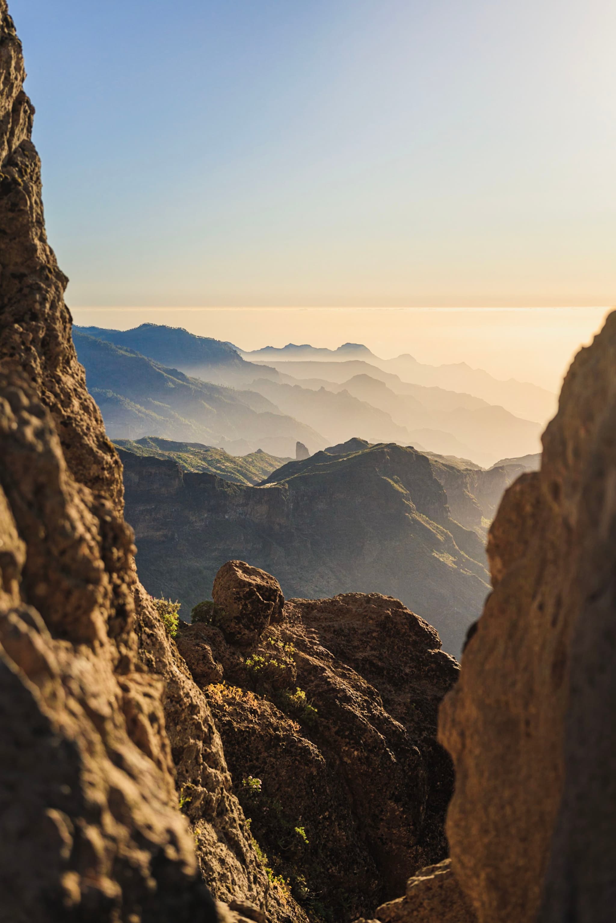 A breathtaking mountain landscape with layers of peaks fading into the distance, framed by rocky cliffs, under a clear blue sky with soft sunlight