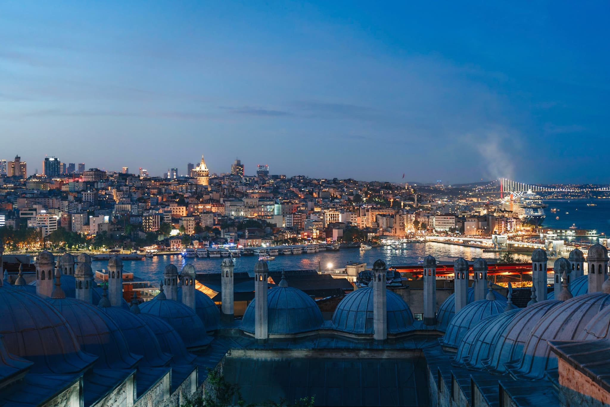 A cityscape at dusk with illuminated buildings, a waterfront, and a view of rooftops in the foreground