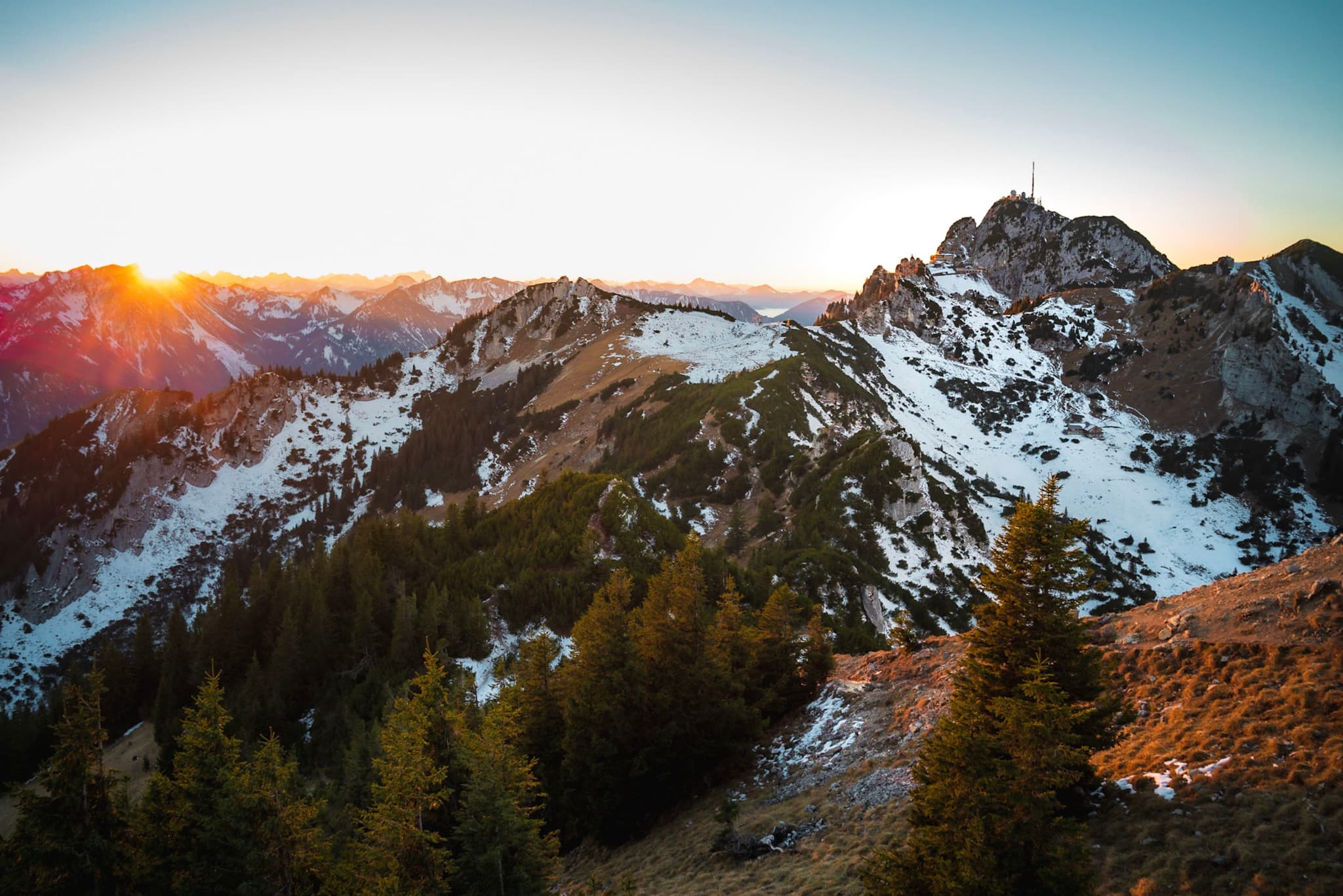 Sunrise over rugged, snow-dusted mountain peaks with evergreen forests in the foreground