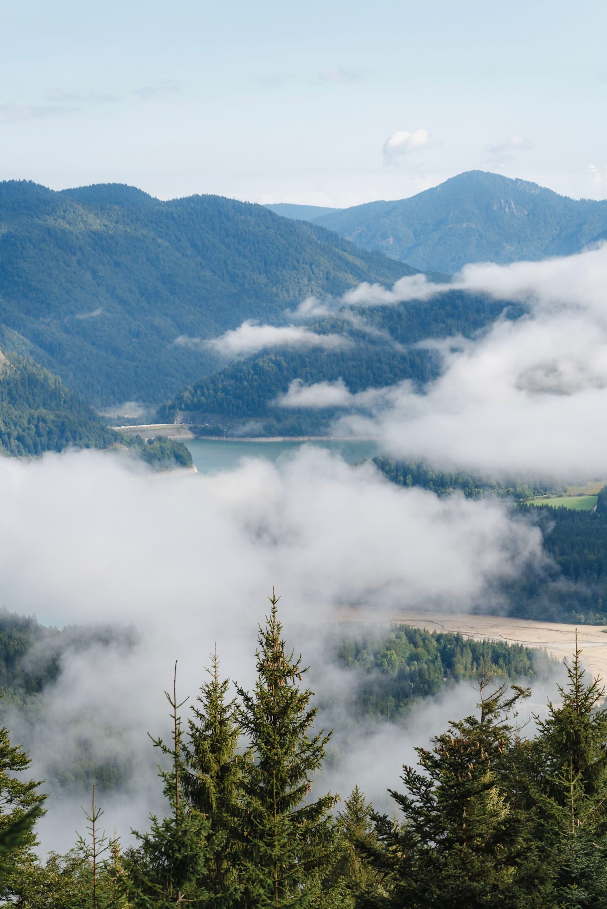 A scenic view of misty mountains with clouds drifting between forested hills and a glimpse of a river or lake in the background