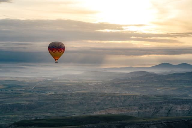 A hot air balloon floats over a vast landscape with rolling hills, under a cloudy sky illuminated by the setting or rising sun