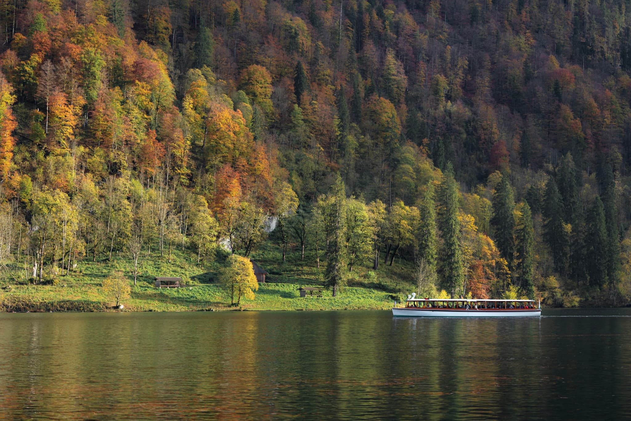 Calm lake with a small boat gliding along a forested autumn hillside reflecting in the water