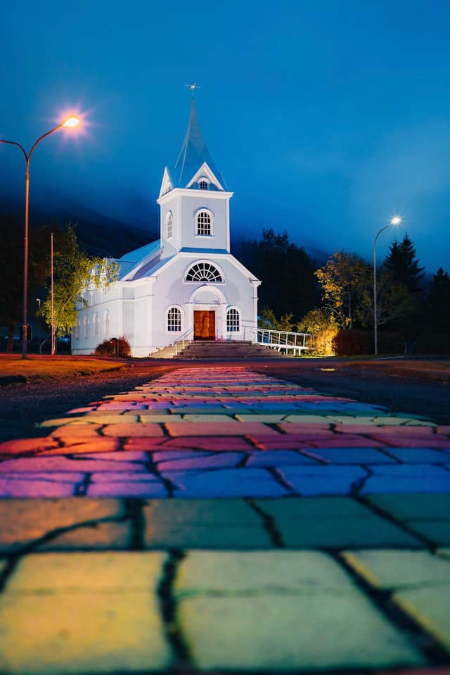 A white church with a steeple is illuminated at night, with a colorful, rainbow-patterned path leading up to it Streetlights and trees are visible in the background