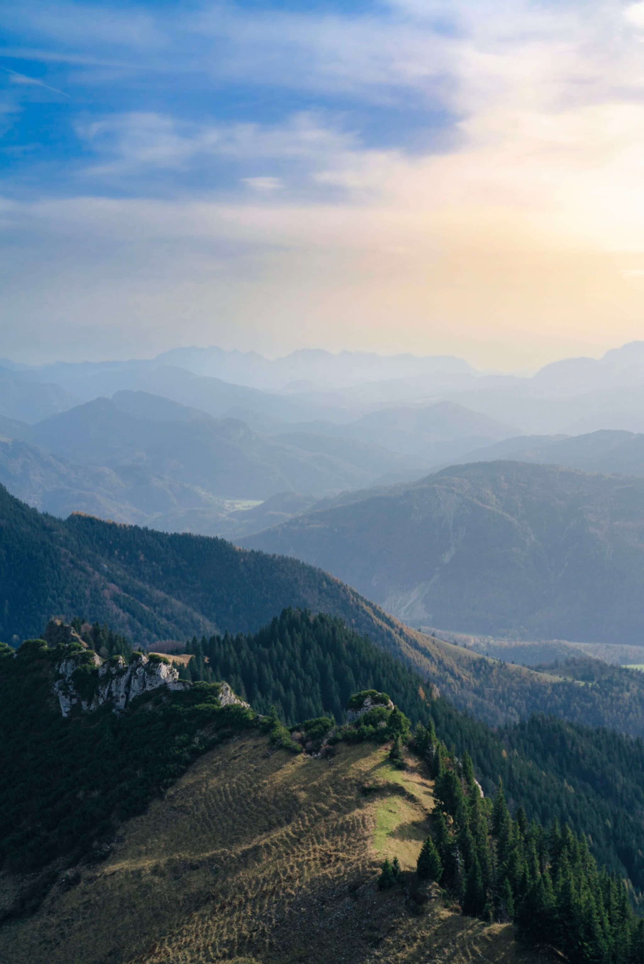 Sunlit mountain ridge with forested slopes descending into hazy blue layers of distant peaks under a soft sky