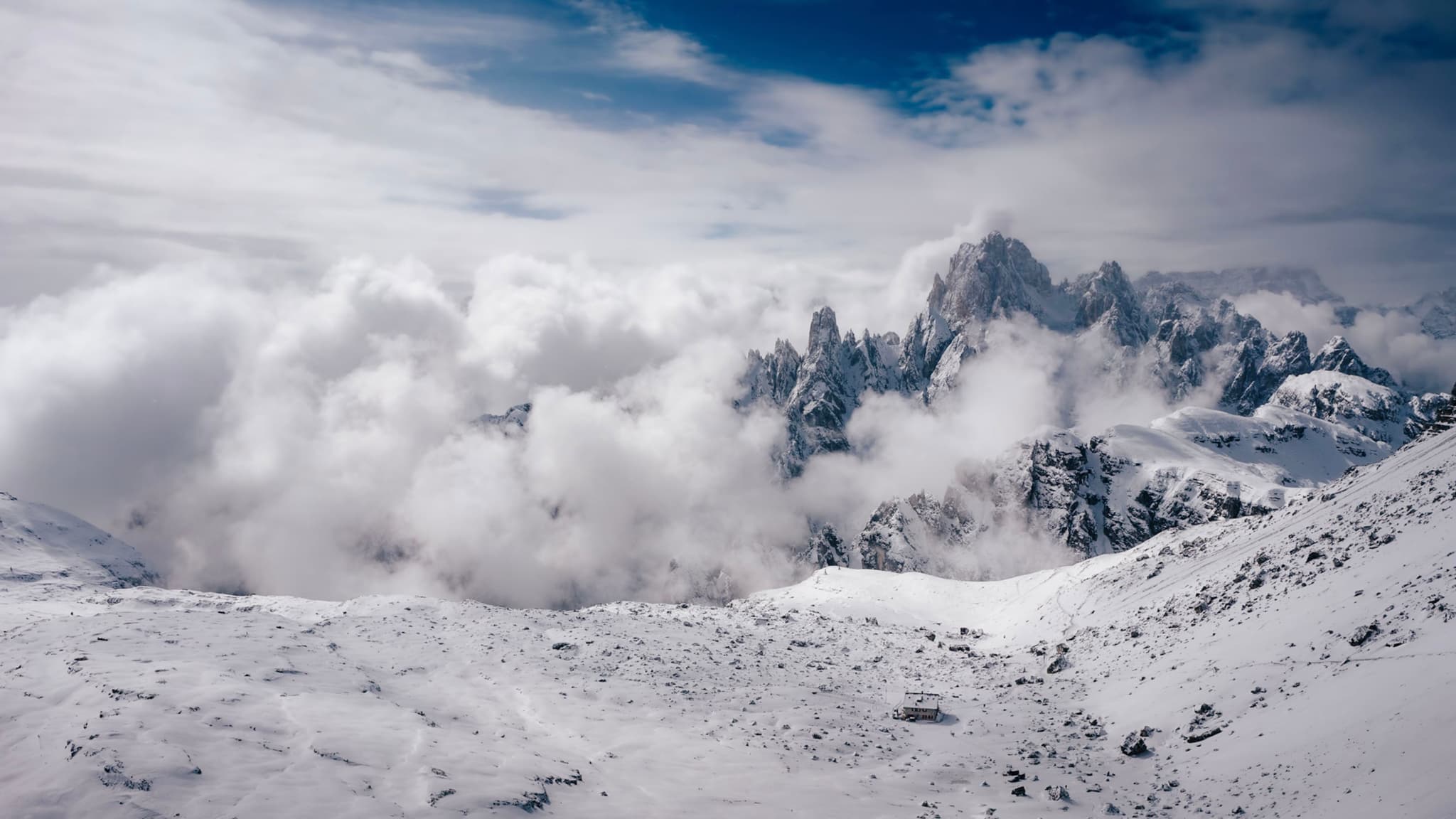 A snowy mountain landscape with rugged peaks and clouds drifting across the scene under a partly cloudy sky