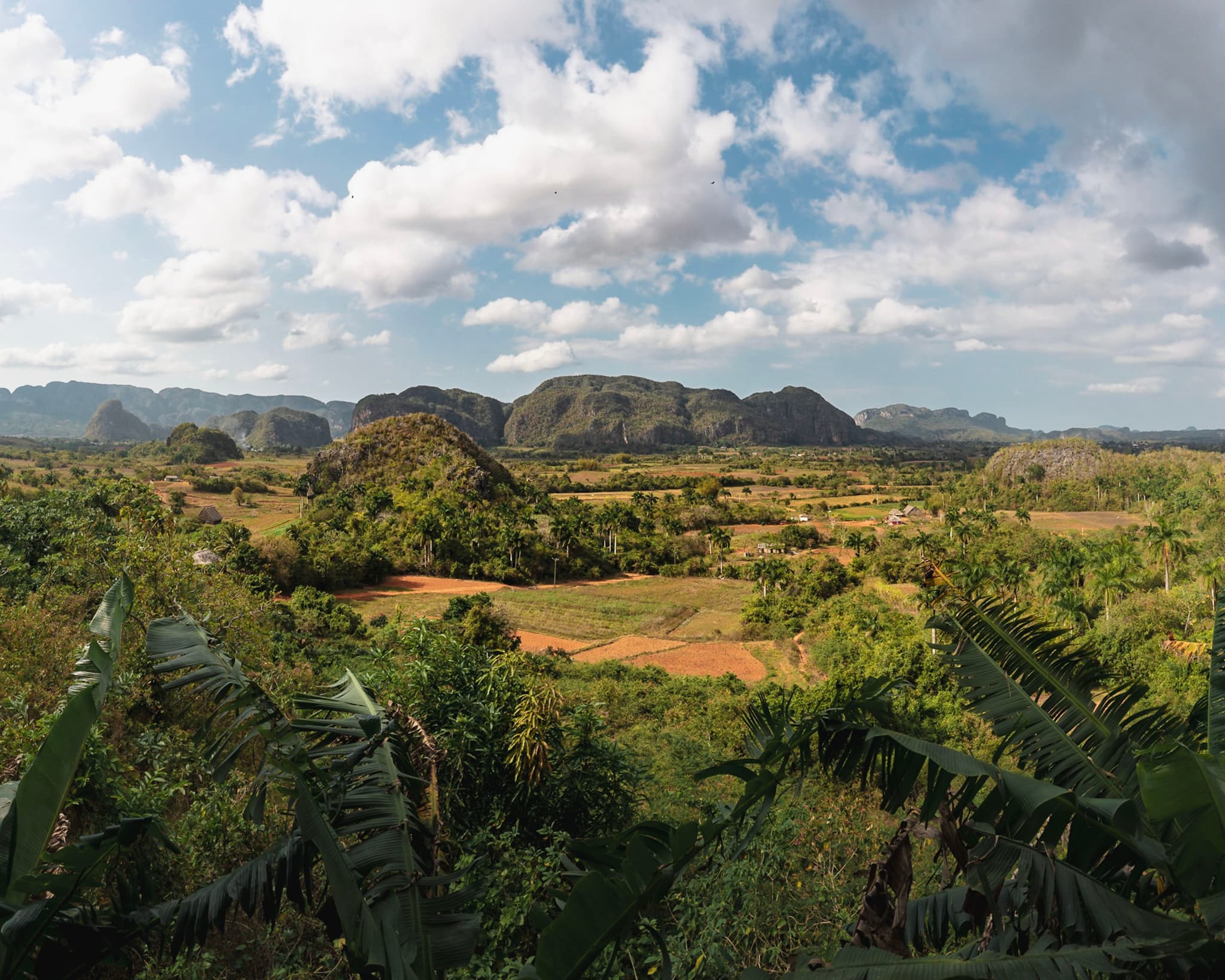 Wide valley with scattered fields and vegetation framed by foreground foliage, backed by rounded mountains under a partly cloudy blue sky