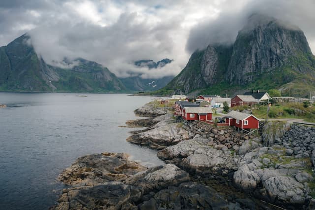 A coastal village with red cabins sits on rocky terrain, surrounded by a calm sea and towering, cloud-covered mountains