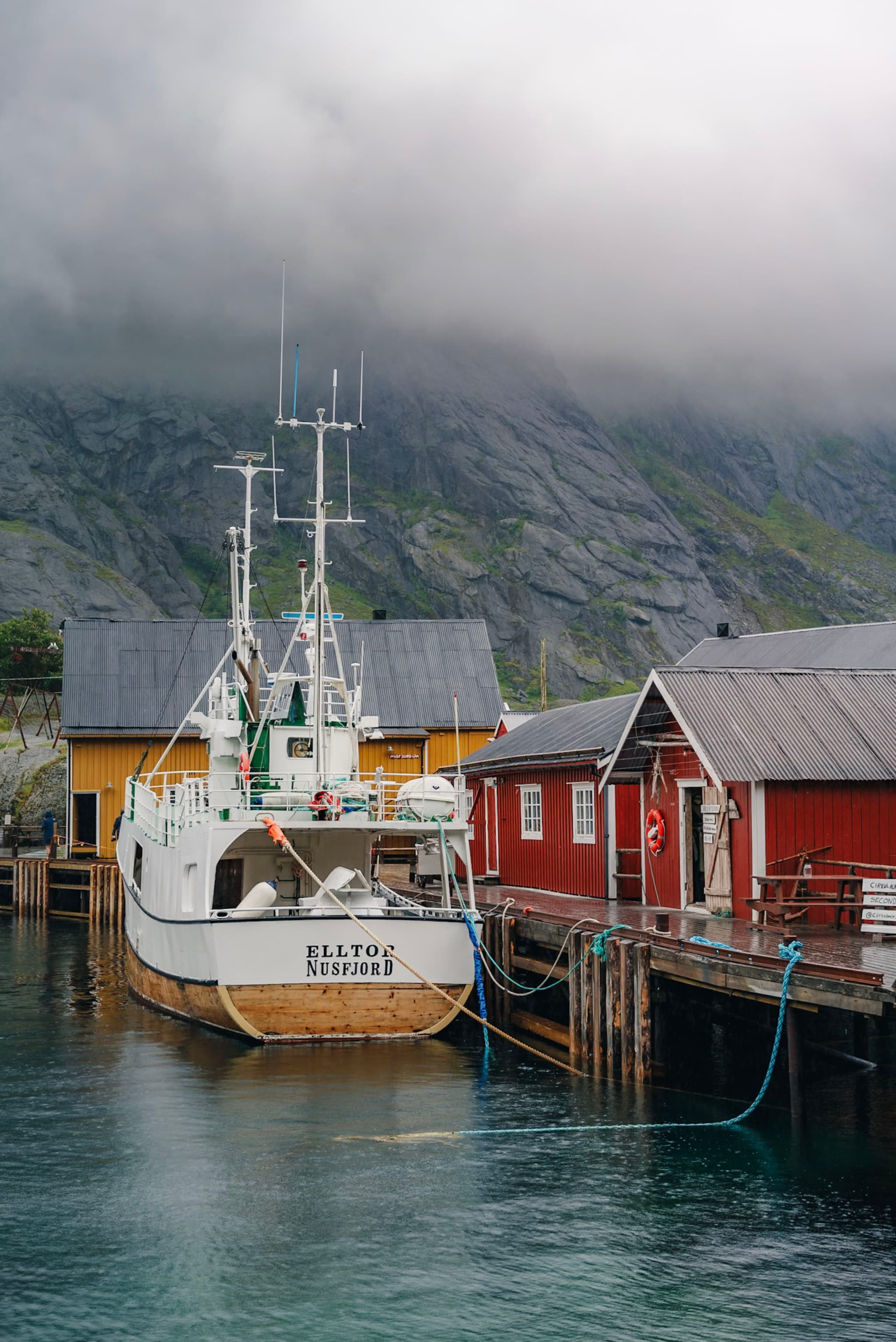 A boat docked at a pier with colorful buildings, surrounded by misty mountains and cloudy skies