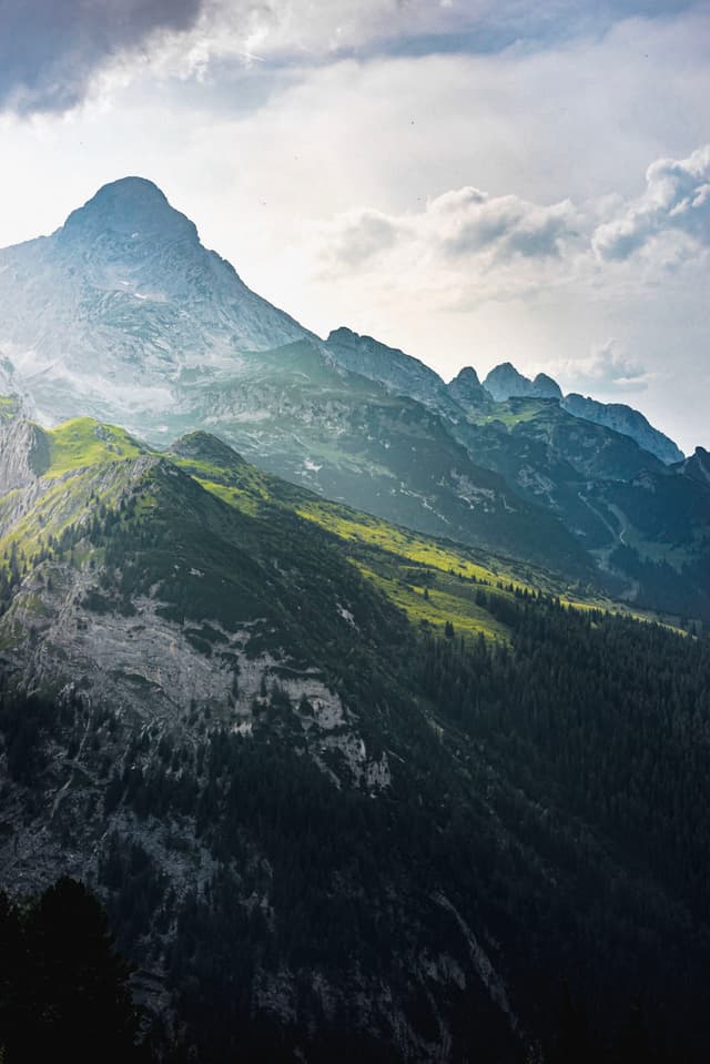 Sunlight slanting across layered mountain ridges with patches of bright green amid darker forested slopes under a cloudy sky