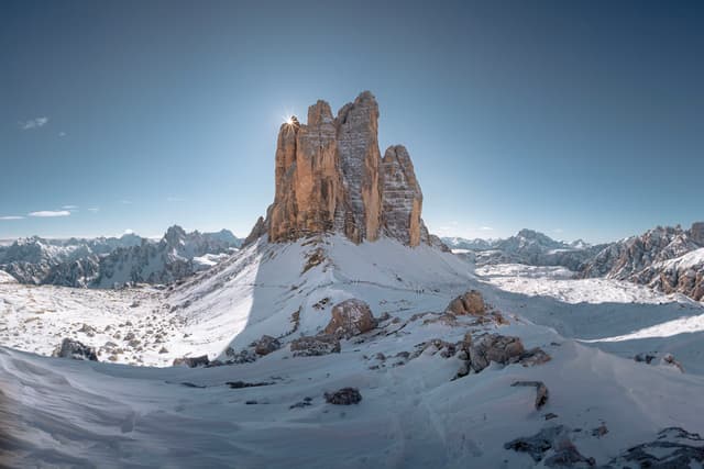 towering rocky peak rises from a vast snow-covered mountain landscape under a clear blue sky