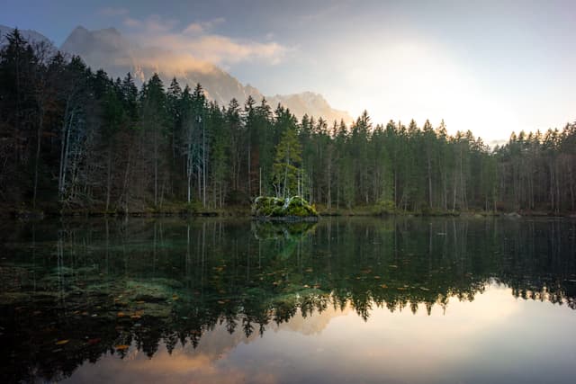 A serene lake reflects a dense forest and distant mountains under a soft, glowing sky