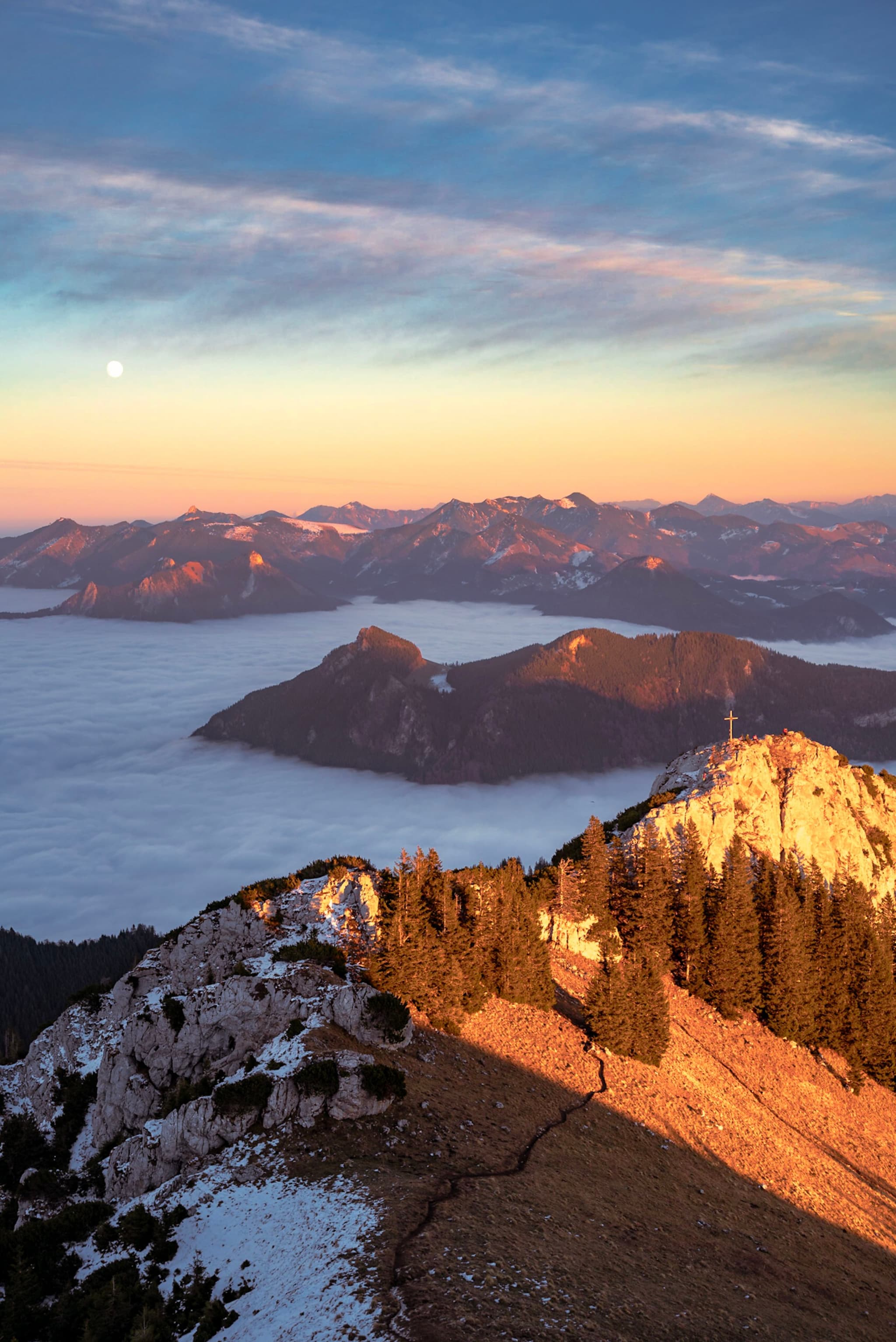 Sunrise light washes over a rocky, partially snow-covered mountain ridge with evergreen trees, overlooking a cloud-filled valley and distant mountain peaks under a pastel sky with a visible moon