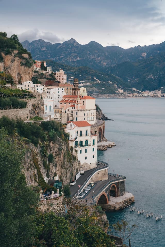 Mediterranean village perched on steep cliffs above a curving coastal road and calm sea with mountains in the distance