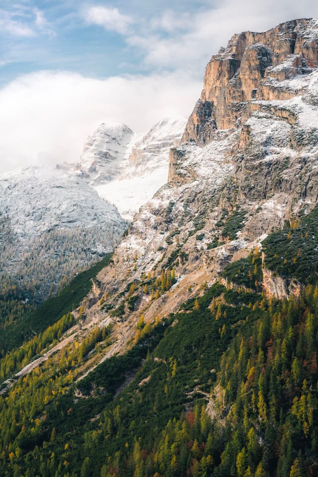 Snow-dusted rocky mountain slope rising above a forest of green and autumn-colored trees under a partly cloudy sky