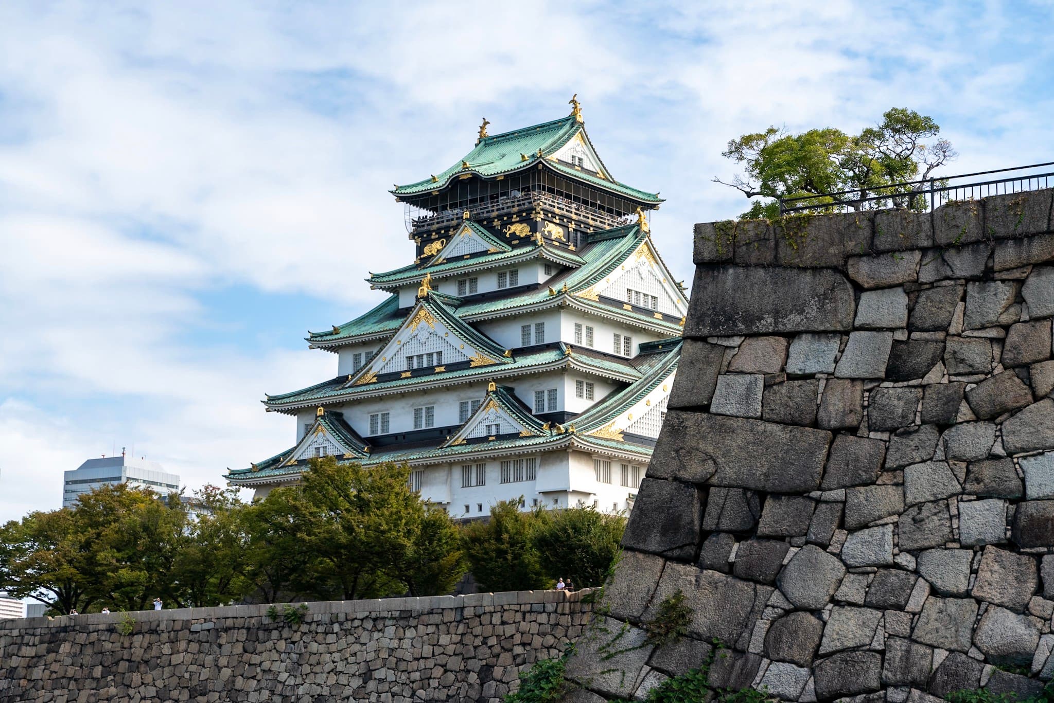 Traditional Japanese castle with green-tiled roofs rising above stone fortification walls and surrounding trees under a partly cloudy sky