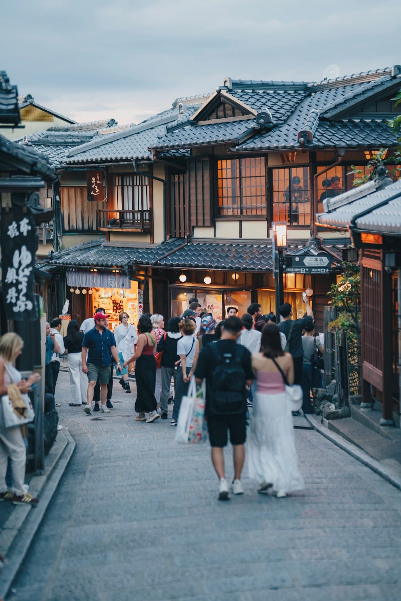 Crowds of people stroll through a narrow traditional Japanese street lined with wooden shops and glowing lanterns at dusk