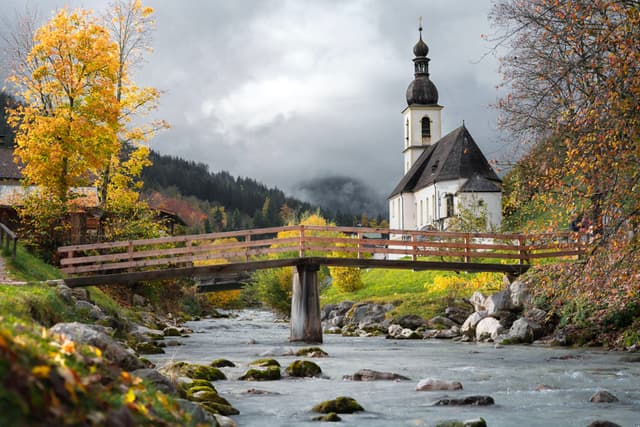 A picturesque scene featuring a church with a tall steeple, a wooden bridge over a flowing river, and autumn trees, set against a backdrop of misty mountains