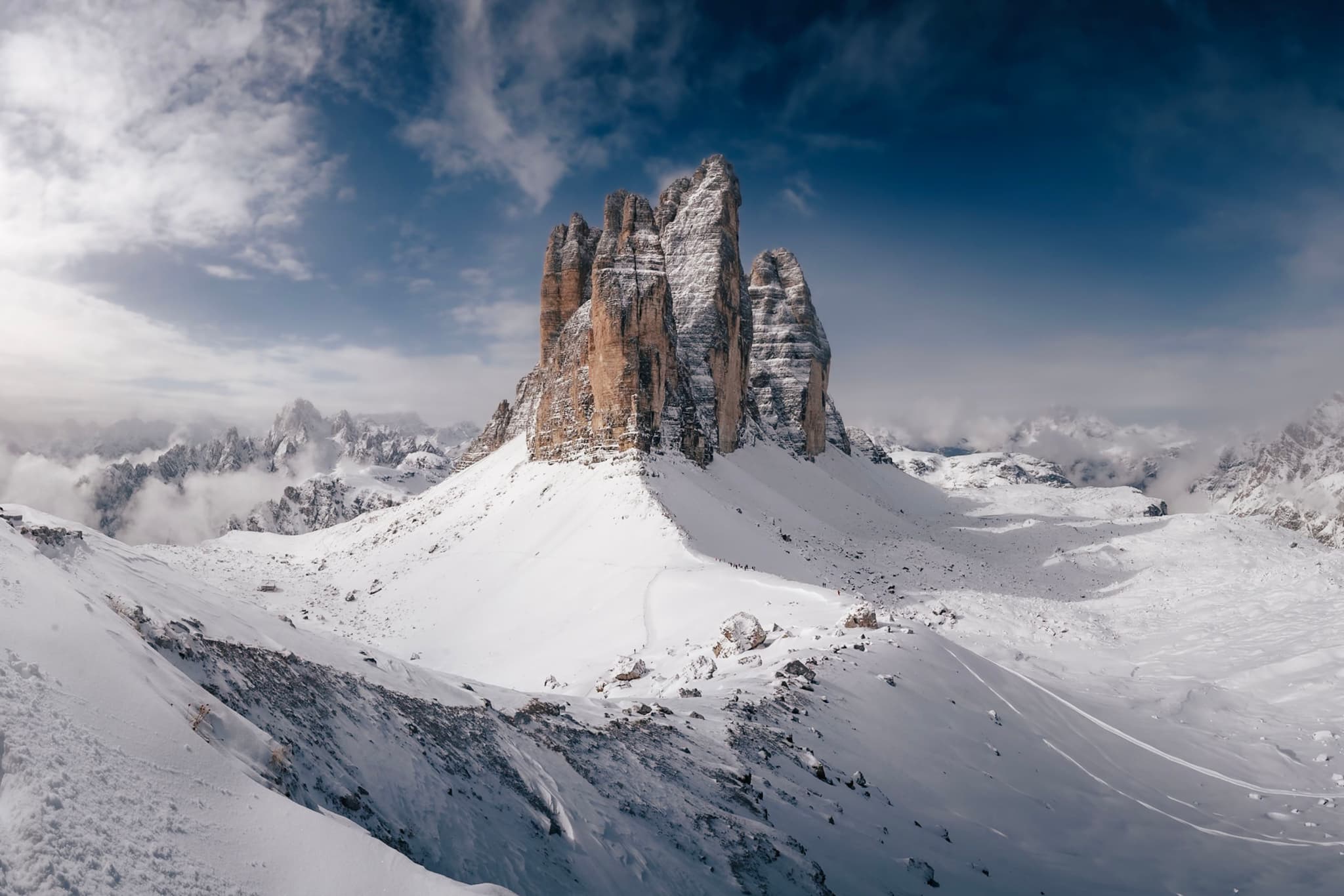 A dramatic mountain peak rises from a snowy landscape under a partly cloudy sky, with rugged terrain and distant mountains in the background