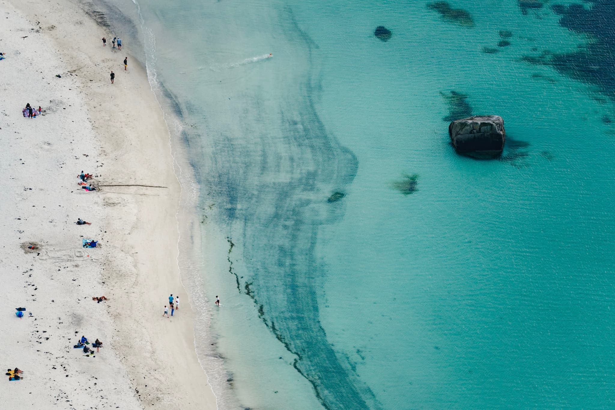 Aerial view of a sandy beach with people relaxing, clear turquoise water, and a partially submerged shipwreck near the shore