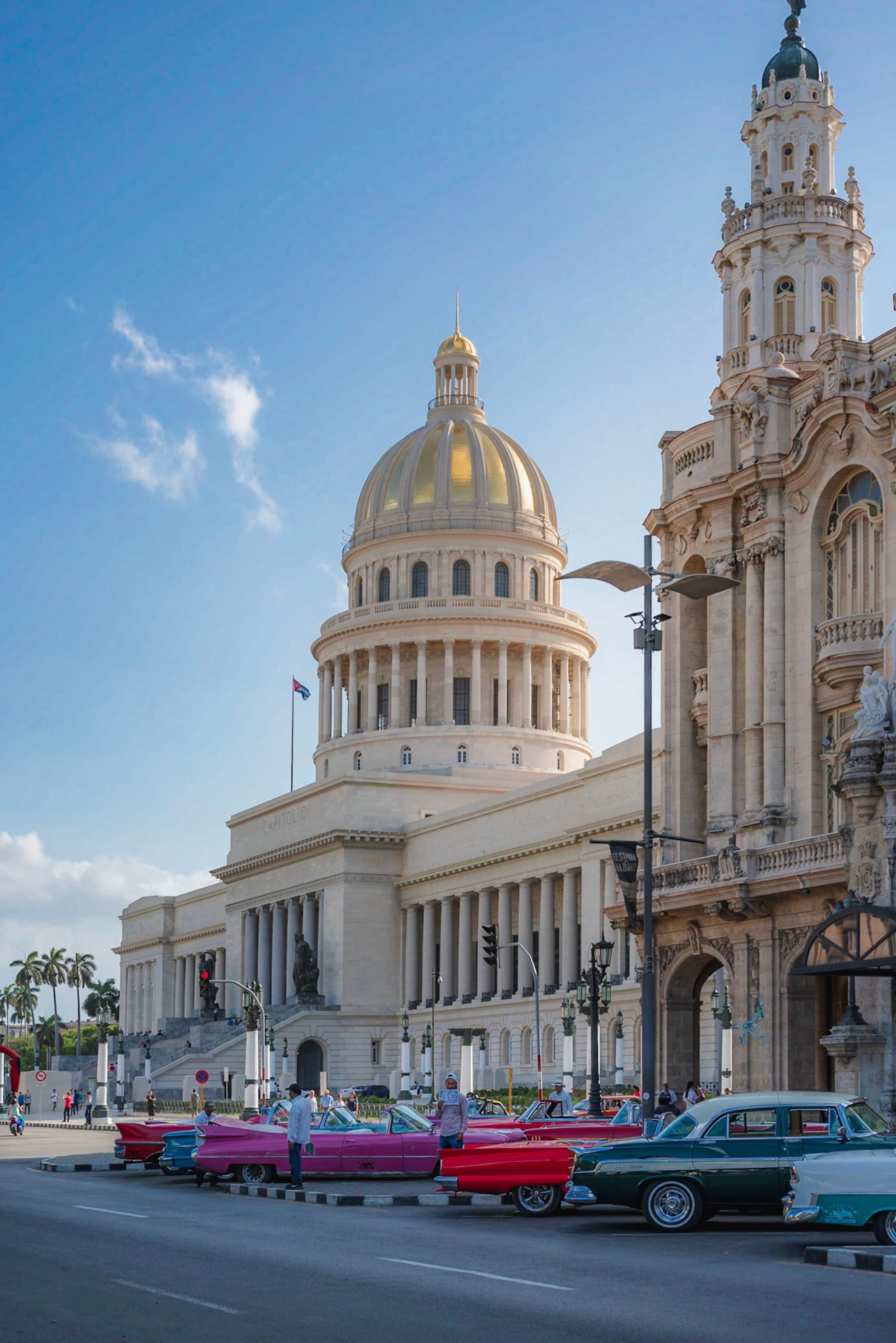 Grand domed capitol building beside ornate historic facade with colorful classic cars lined up along the city street under a bright blue sky