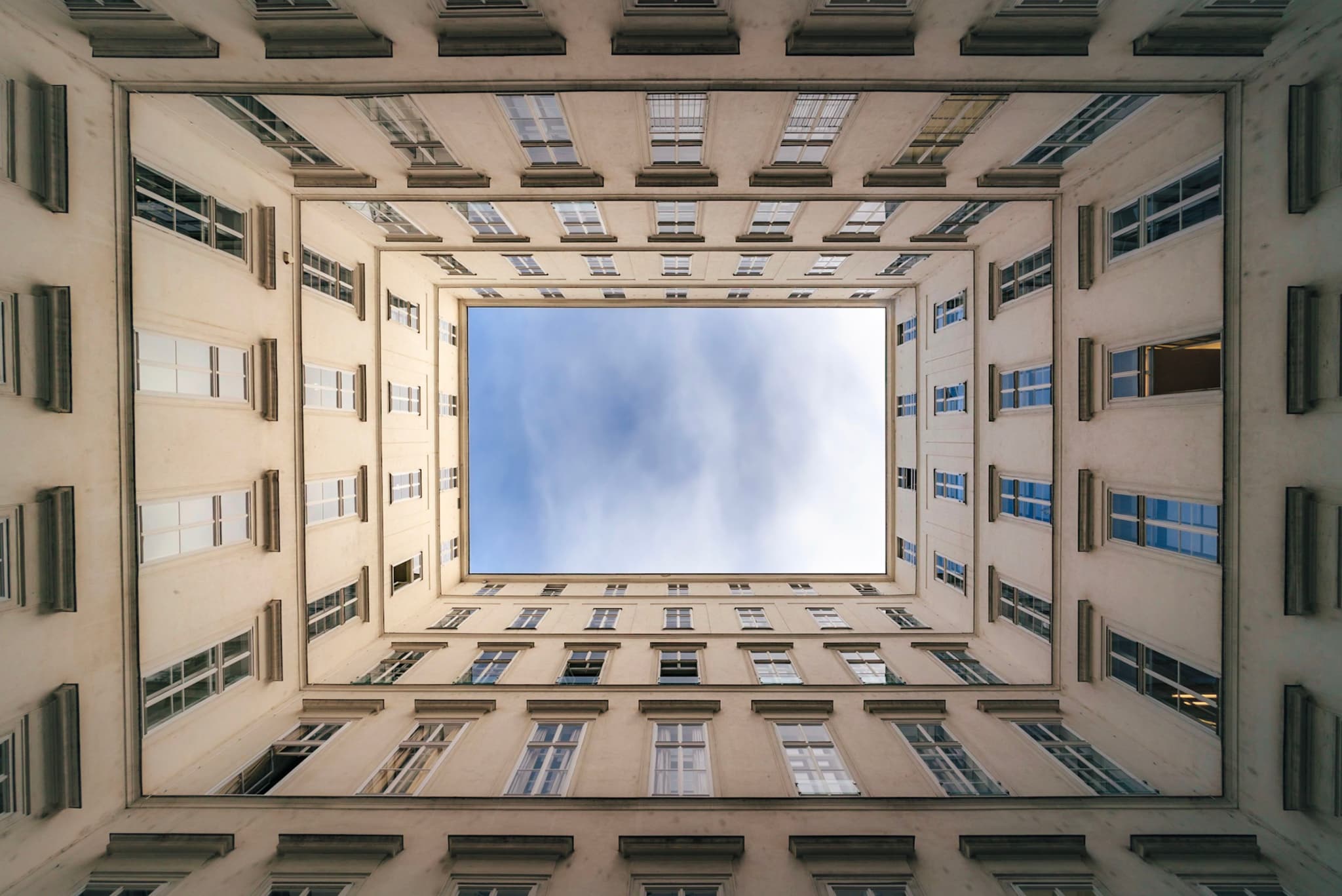 Symmetrical view looking straight up from the center of a rectangular courtyard, with surrounding walls and windows forming a frame around a patch of blue sky and clouds