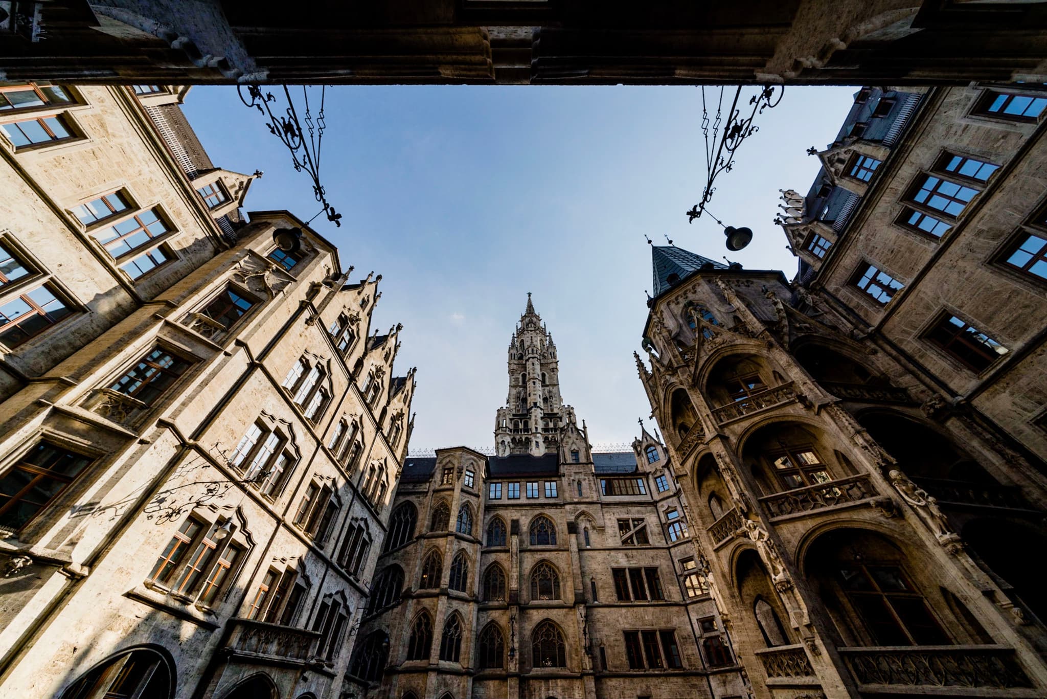 Upward view from a narrow courtyard framed by ornate historic stone buildings toward a central gothic-style tower against clear blue sky
