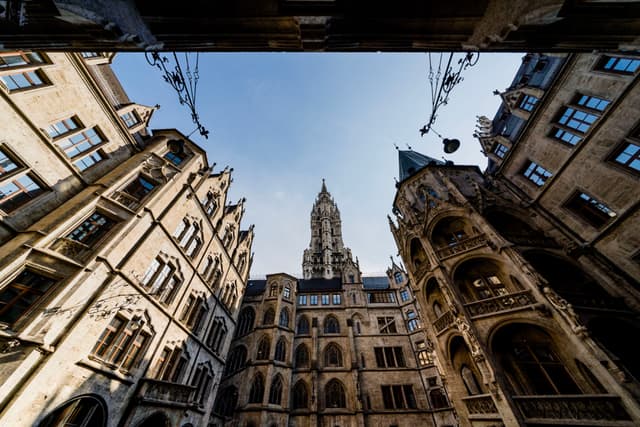 Upward view from a narrow courtyard framed by ornate historic stone buildings toward a central gothic-style tower against clear blue sky