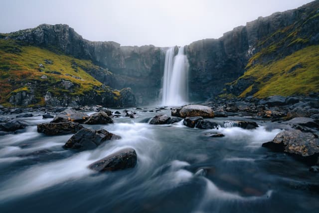 Waterfall pouring over a rocky cliff into a smooth, flowing stream surrounded by mossy slopes under an overcast sky
