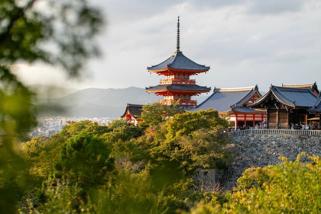Traditional Japanese pagoda and temple buildings on a tree-covered hill overlooking a distant cityscape at sunset