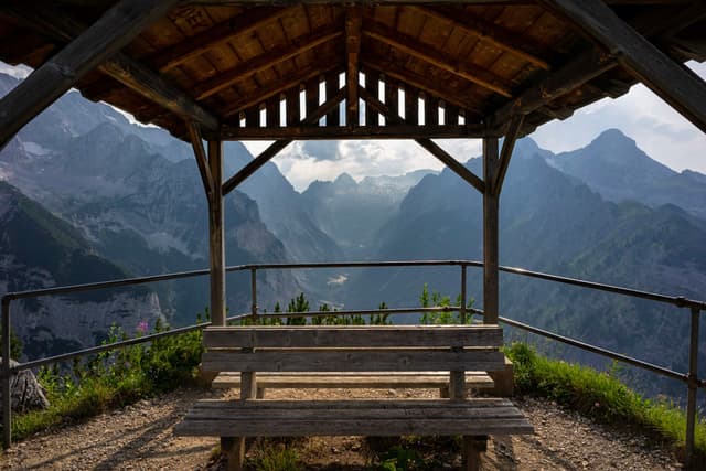 A wooden shelter with a picnic table overlooks a scenic mountain landscape, framed by a railing and surrounded by lush greenery