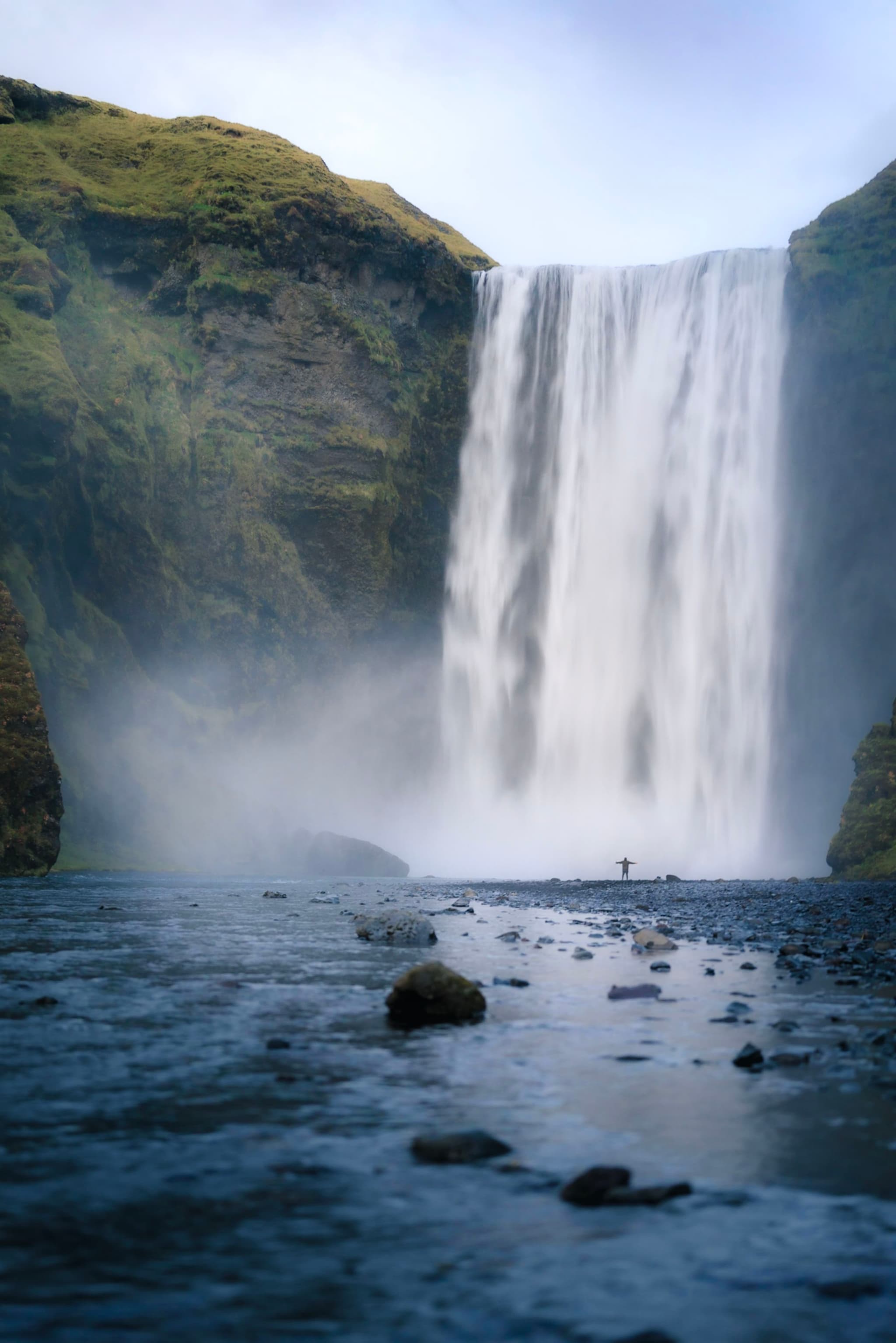 towering waterfall plunging into a misty river valley with a lone person standing near the base